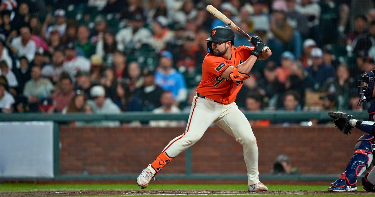 Drew Cavanaugh stands in the batter’s box with a bat raised, preparing to swing during a baseball game. Wearing an orange jersey and white pants, Cavanaugh faces the pitcher as a catcher crouches behind home plate, with a stadium crowd visible in the background.